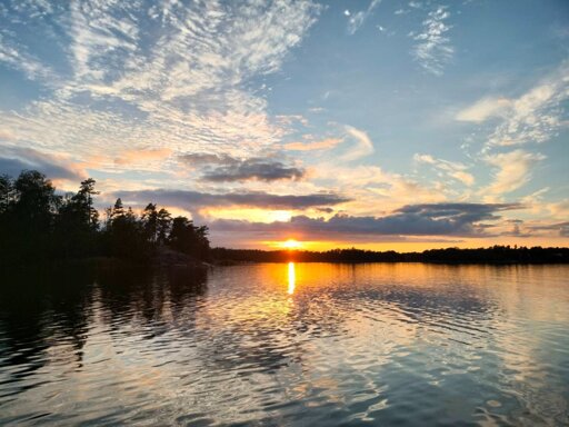 Photo of a sunset partially blocked by clouds over water and a dark forest.
